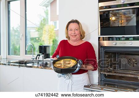 Stock Image - Portrait of senior woman with cake in kitchen. Fotosearch