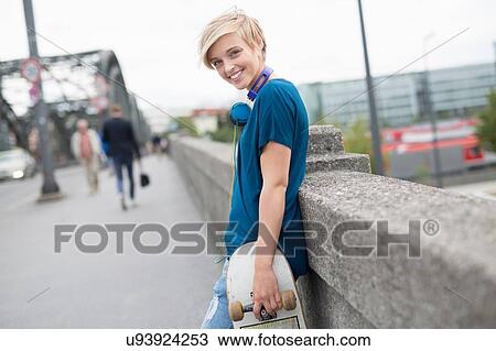 Stock Image - Portrait of young woman holding skateboard on bridge. Fotosearch