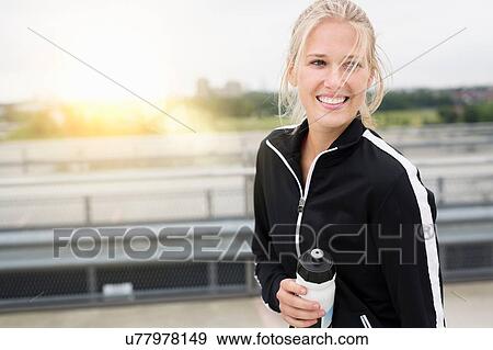 Stock Photo - Portrait of young woman, wearing sports clothing, holding water bottle, outdoors. Fotosearch