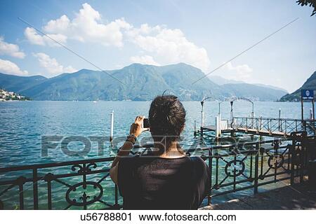 Stock Image - Rear view of mid adult woman photographing with smartphone, Lake Lugano, Switzerland. Fotosearch