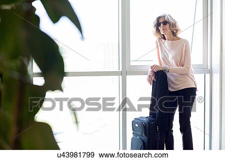 Stock Photo - Senior businesswoman waiting with wheeled suitcase in office building. Fotosearch