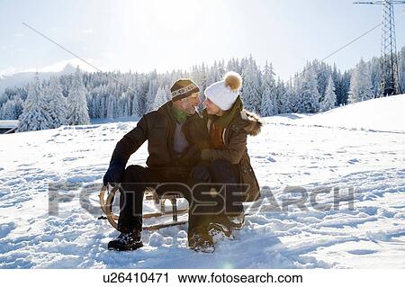 Stock Image - Senior couple on snowy landscape sitting on sledge face to face smiling, Sattelbergalm, Tyrol, Austria. Fotosearch