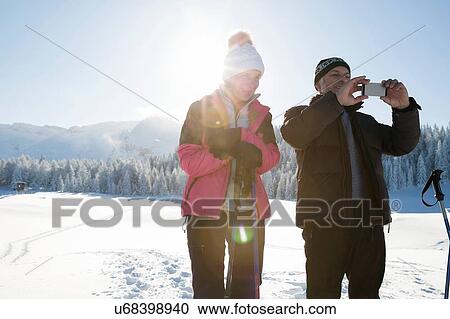 Stock Image - Senior couple on snowy landscape using smartphone to take photograph, Sattelbergalm, Tyrol, Austria. Fotosearch