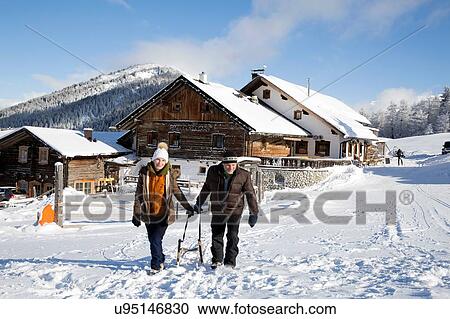 Stock Image - Senior couple pulling sledge and log cabin, Sattelbergalm, Tyrol, Austria. Fotosearch