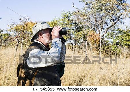Stock Photography - Senior man looking through binoculars on safari, Kafue National Park, Zambia. Fotosearch