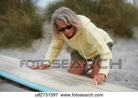 Stock Photo - Senior woman on beach, waxing surfboard. Fotosearch