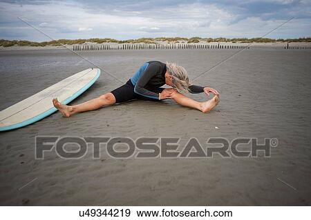 Stock Photo - Senior woman sitting on sand, stretching, surfboard beside her. Fotosearch