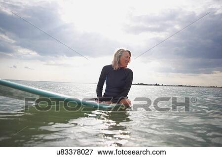 Stock Photo - Senior woman sitting on surfboard in sea. Fotosearch