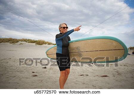 Stock Image - Senior woman standing on beach, holding surfboard, pointing to sea. Fotosearch