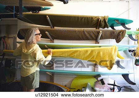 Picture - Senior woman taking surfboard from shelf. Fotosearch