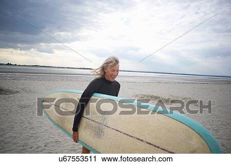 Stock Image - Senior woman walking from sea, carrying surboard. Fotosearch