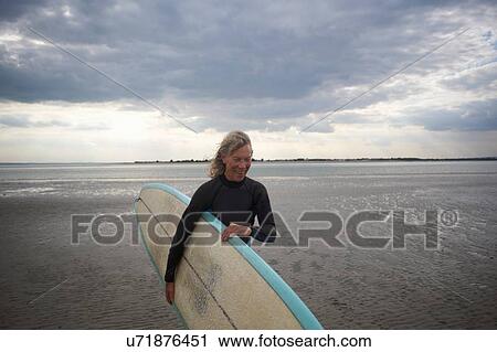 Stock Image - Senior woman walking from sea, carrying surfboard. Fotosearch