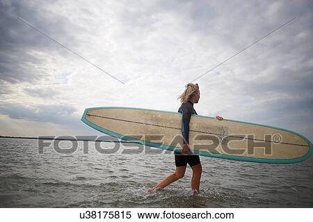 Stock Photography - Senior woman walking from sea, carrying surboard. Fotosearch