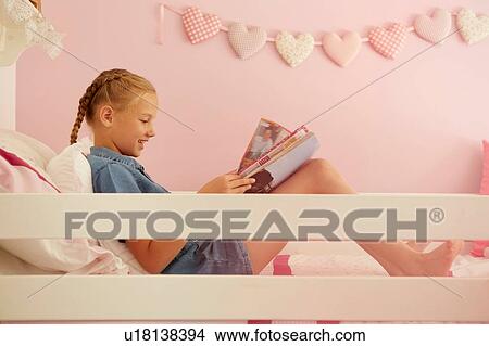 Picture - Side view of girl sitting on bed reading magazine. Fotosearch