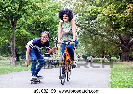 Stock Photo - Skateboarding boy holding onto mothers bicycle in park. Fotosearch