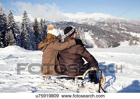 Stock Photo - Snow capped mountains and rear view of senior couple sitting on sledge looking away, Sattelbergalm, Tyrol, Austria. Fotosearch