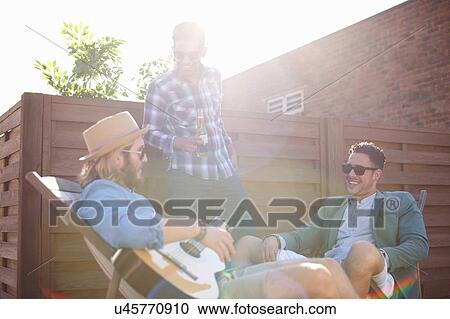 Three male friends chatting and playing guitar at rooftop party View Large Photo Image Stock Image - Three male friends chatting and playing guitar at rooftop party. Fotosearch