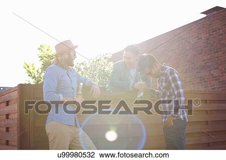 Stock Image - Three male friends chatting and drinking beer at rooftop party. Fotosearch
