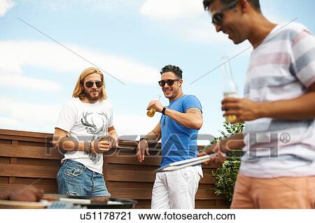 Stock Image - Three male friends drinking beer and barbecuing at rooftop barbecue. Fotosearch