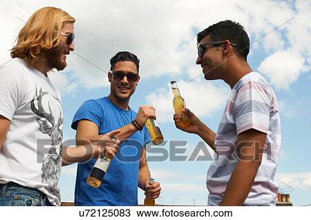 Stock Image - Three male friends drinking beer at rooftop party. Fotosearch