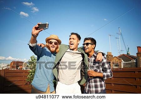 Stock Photo - Three male friends taking camera selfie at rooftop party. Fotosearch