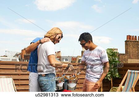 Stock Image - Three male friends with beer bottles and ice buckets at rooftop barbecue. Fotosearch