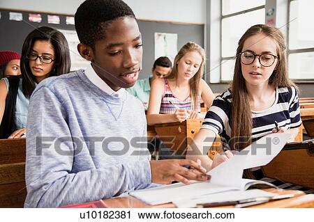 Stock Image - Two students sitting at desk in classroom, helping each other with work. Fotosearch