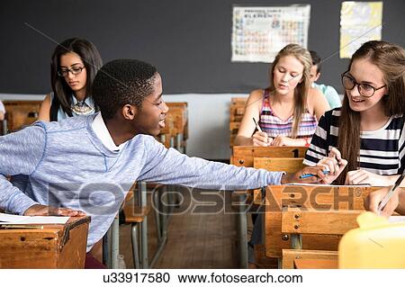 Stock Image - Two students sitting at desk in classroom, helping each other with work. Fotosearch