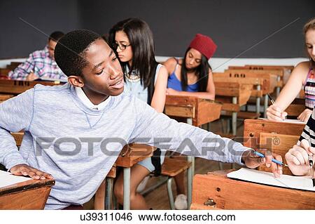 Stock Photograph - Two students sitting at desk in classroom, helping each other with work. Fotosearch