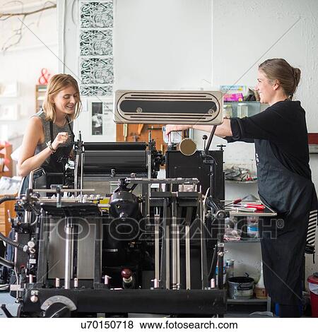 Stock Photo - Two women working with traditional letterpress print machine in workshop. Fotosearch