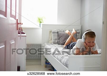 View through door of boy lying on bed wearing headphones looking down on digital tablet View Large Photo Image Stock Photo - View through door of boy lying on bed wearing headphones looking down on digital tablet. Fotosearch