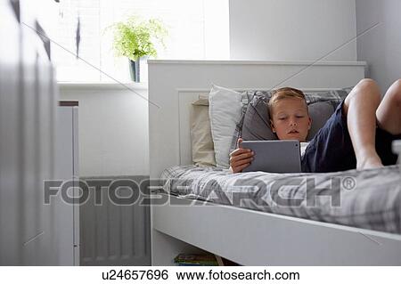 Stock Photograph - View through doorway of boy lying on bed looking down at digital tablet. Fotosearch