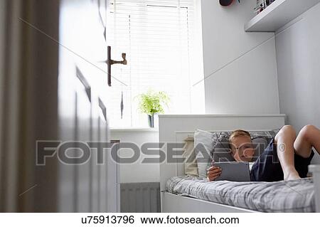 Stock Photograph - View through doorway of boy lying on bed looking down at digital tablet. Fotosearch
