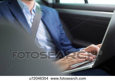 Picture - Young businessman chatting typing on laptop in back seat of car. Fotosearch