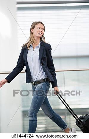 Picture - Young businesswoman walking at airport with wheeled suitcase. Fotosearch