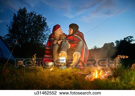 Stock Image - Young camping couple sitting by campfire at dusk. Fotosearch