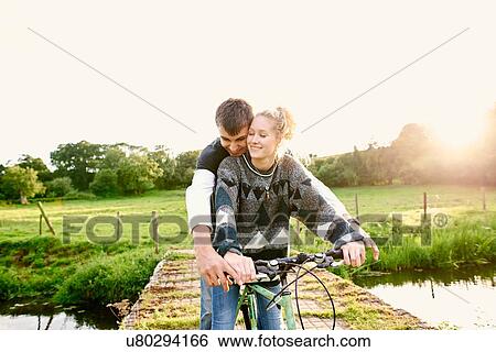 Stock Photograph - Young couple sharing bicycle at river footbridge. Fotosearch