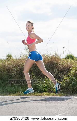 Stock Photograph - Young female runner looking over her shoulder whilst running on rural road. Fotosearch