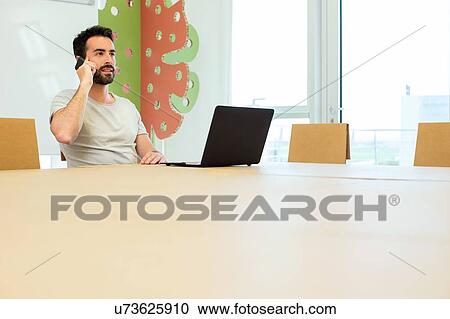 Stock Image - Young male designer talking on smartphone at boardroom table. Fotosearch