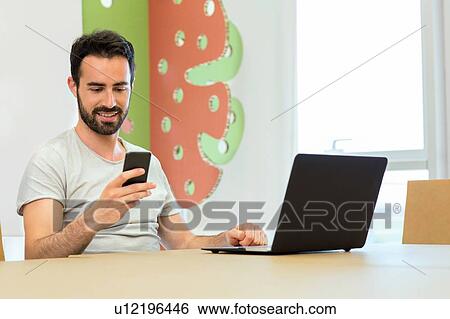 Stock Photograph - Young male designer texting on smartphone at boardroom table. Fotosearch