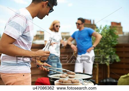 Young man barecuing burgers at rooftop barbecue View Large Photo Image Stock Image - Young man barecuing burgers at rooftop barbecue. Fotosearch
