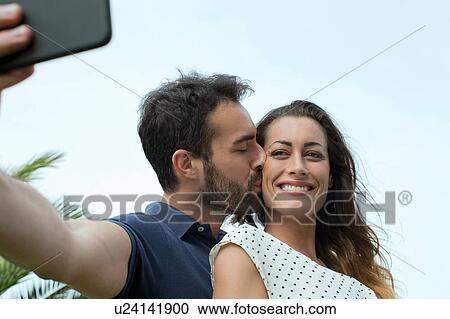 Stock Image - Young man kissing girlfriend on cheek for smartphone selfie. Fotosearch