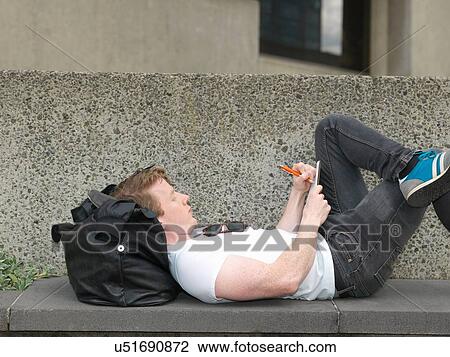 Stock Image - Young man lying on concrete bench, writing notes. Fotosearch