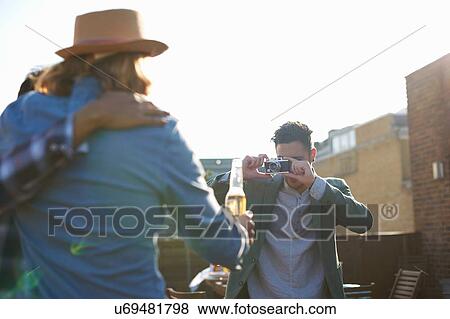 Stock Photo - Young man photographing male friends at rooftop party. Fotosearch