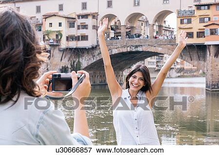Stock Photo - Young woman arms raised posing for photograph in front of Ponte Vecchio and river Arno, Florence, Tuscany, Italy. Fotosearch