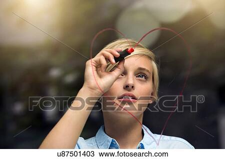 Stock Image - Young woman drawing lipstick heart shape on window. Fotosearch