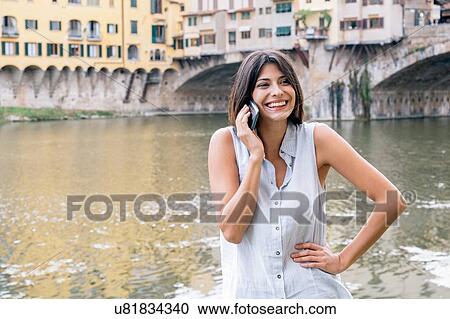 Stock Image - Young woman hand on hip, talking on cellular phone in front of Ponte Vecchio and river Arno looking away smiling, Florence, Tuscany, Italy. Fotosearch