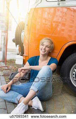 Stock Image - Young woman sitting cross legged leaning against campervan. Fotosearch