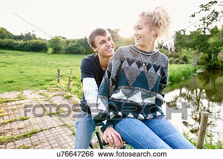 Stock Photograph - Young woman sitting on boyfriends bicycle handlebars at river footbridge. Fotosearch
