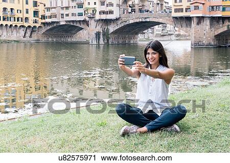 Young woman sitting on riverbank in front of Ponte Vecchio and Arno river using smartphone to take selfie, Florence, Tuscany, Italy View Large Photo Image Stock Image - Young woman sitting on riverbank in front of Ponte Vecchio and Arno river using smartphone to take selfie, Florence, Tuscany, Italy. Fotosearch